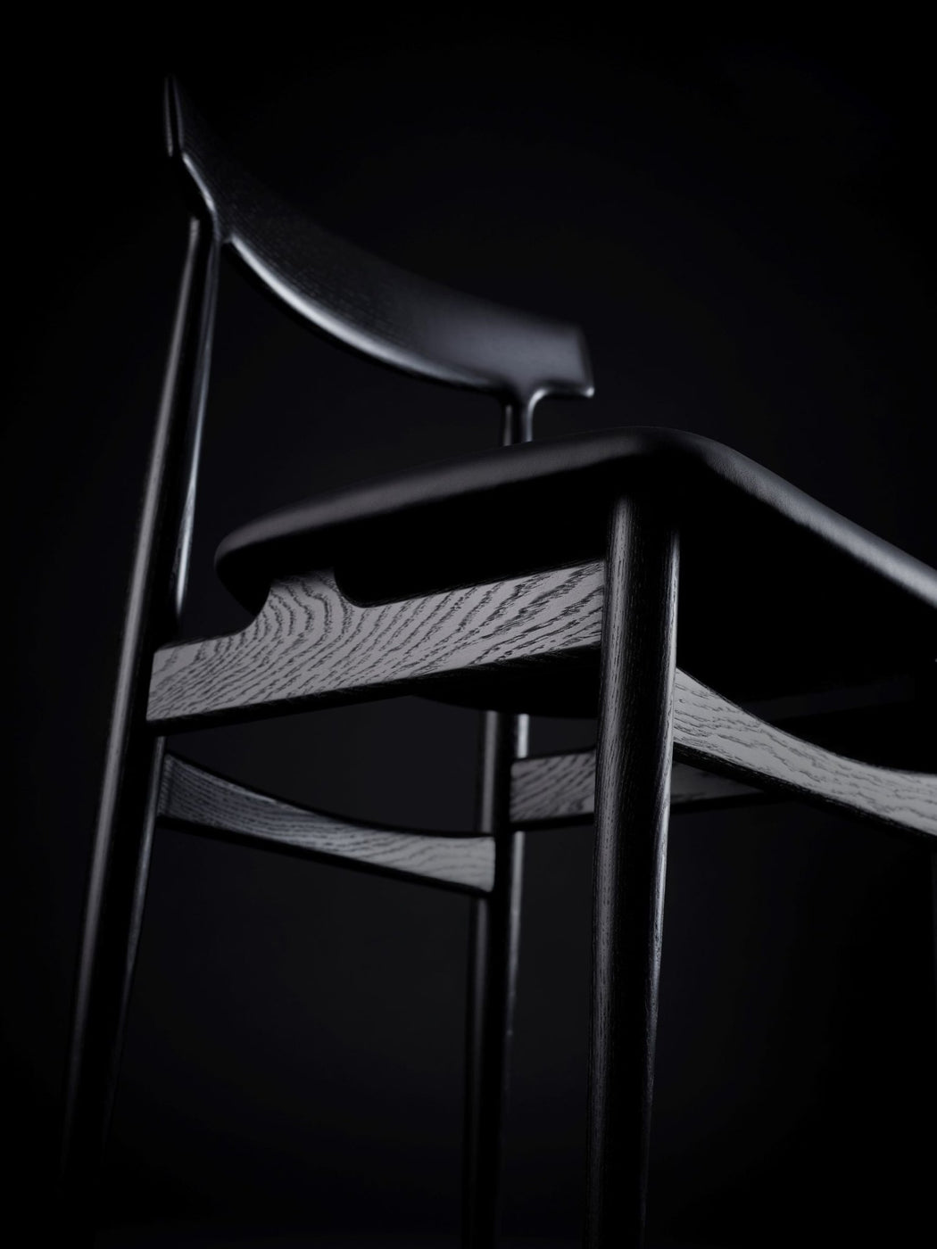 Close-up of a wooden chair with black seat cushion against a dark background, highlighting wood grain and smooth finish.